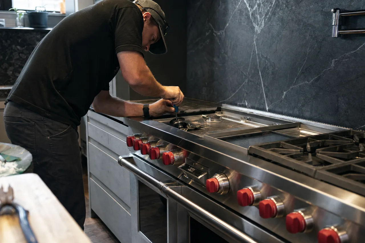 Technician crouched at a stainless dual-fuel range, one burner grate lifted aside, checking the igniter with a hand tool