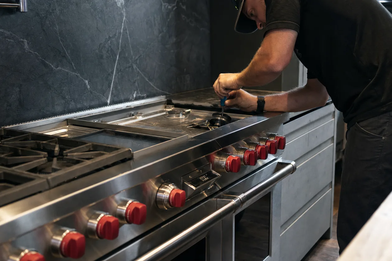 A technician leans over a wide stainless range with red knobs, one burner grate lifted away while he works a small screwdriver at the burner base, a dark veined stone backsplash behind